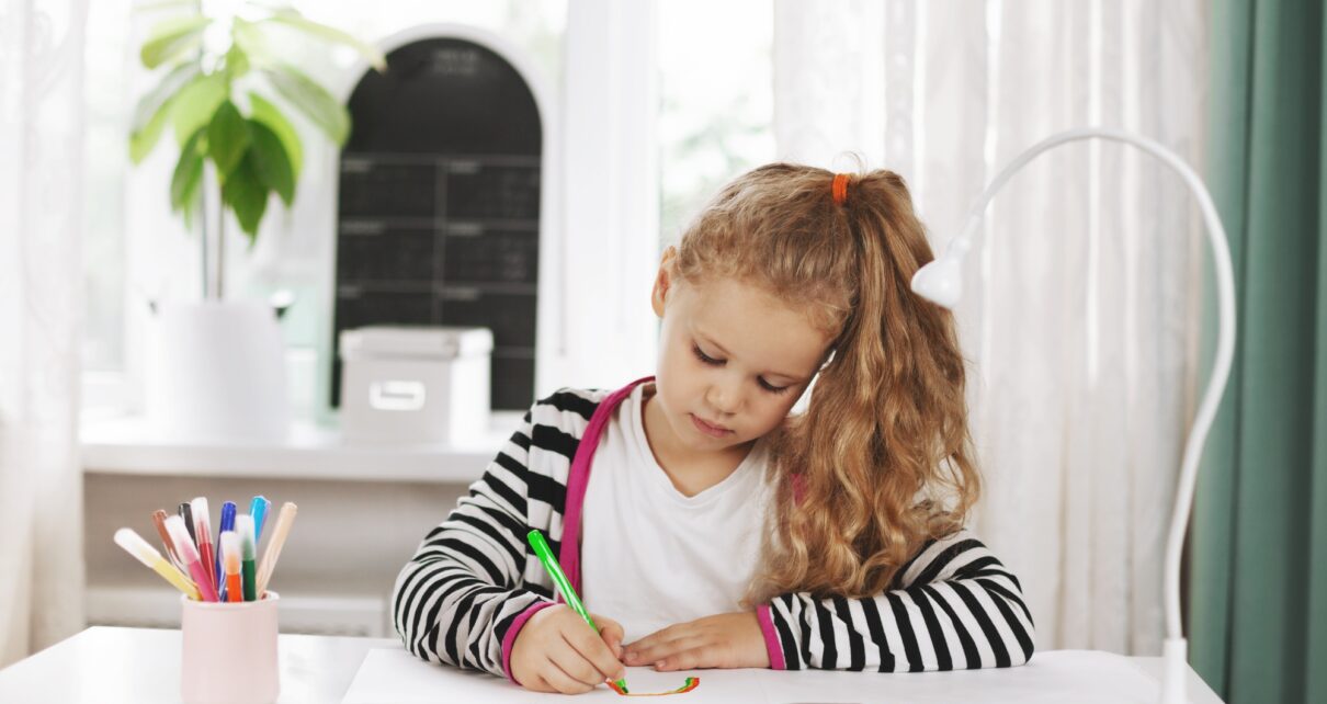 A charming girl sits at a table and draws a rainbow in an album with colored pencils.