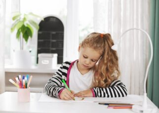 A charming girl sits at a table and draws a rainbow in an album with colored pencils.