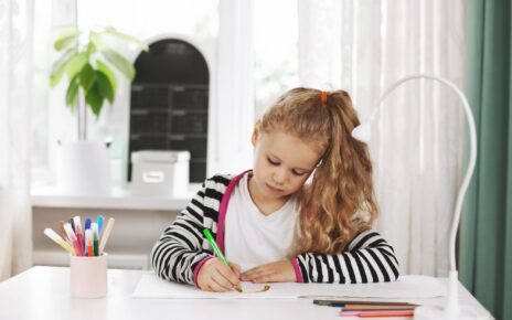 A charming girl sits at a table and draws a rainbow in an album with colored pencils.